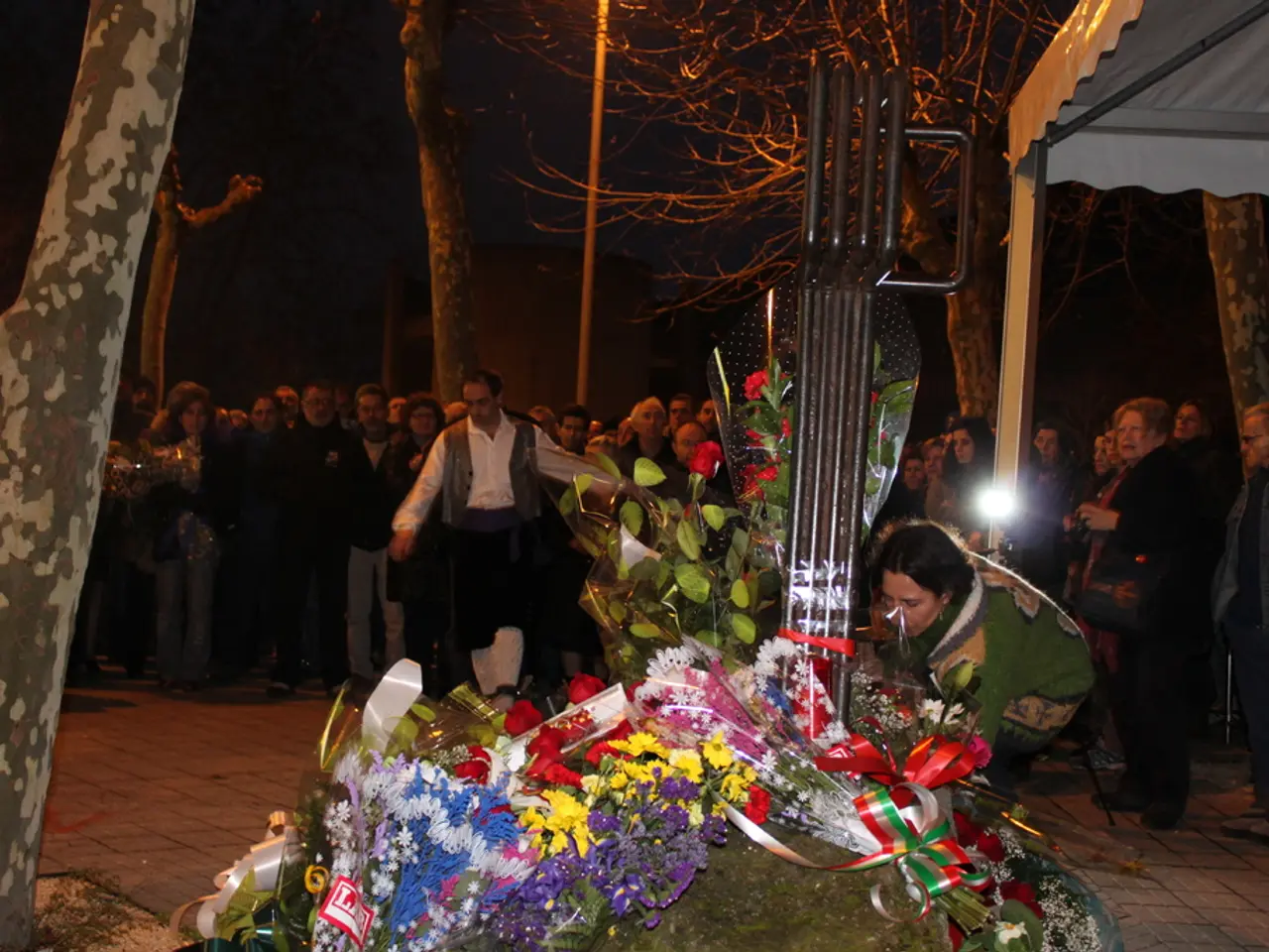 In the image we can see there are lot of people standing and there are flower bouquets kept on the...