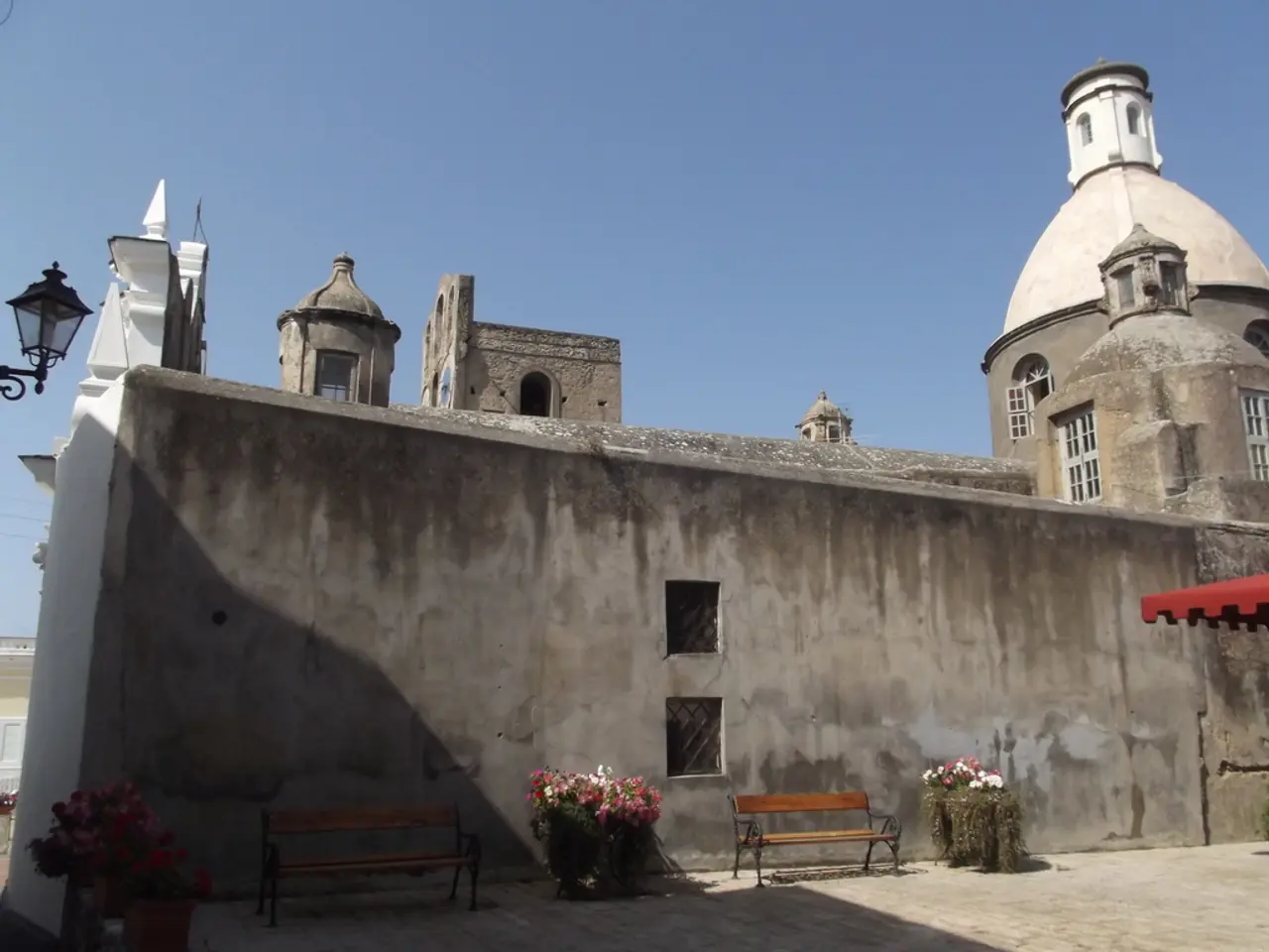 Here we can see a building,windows and at the wall there are benches and house plants. On the left...