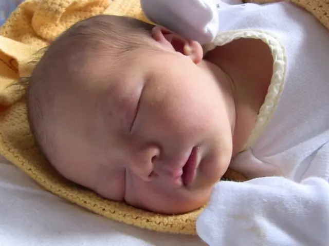 In this image a baby is wearing a white dress is lying on the cloth. Baby is wearing gloves.